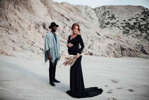 pareja multiétnica posando contra el cañón de granito. Mujer embarazada vistiendo vestido negro con estilo  - Foto, Imagen
