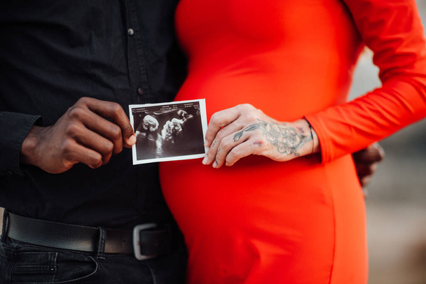  couple holding ultrasound photo in hands. Pregnant woman wearing stylish red dress  - Photo, Image