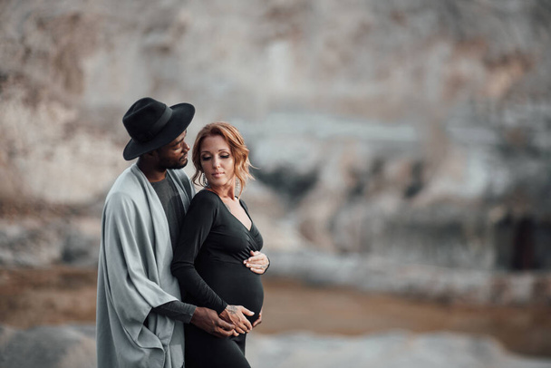 africano americano hombre abrazando embarazada mujer en negro vestido, pareja posando contra granito cañón - Foto, imagen