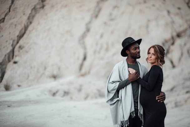 bel homme afro-américain étreignant femme enceinte en robe noire, couple posant contre canyon de granit - Photo, image