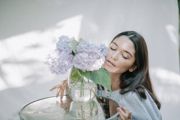 Девушка касаясь гортензии цветы в вазе на декоративном стеклянном столе   - Фото, изображение