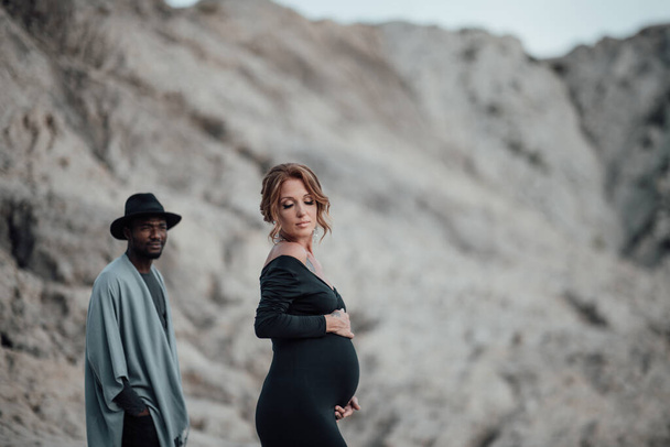 multi ethnic couple posing against granite canyon. Pregnant woman wearing stylish black dress  - Photo, Image