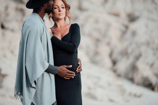 mujer embarazada en vestido negro abrazando hombre afroamericano, pareja posando contra cañón de granito - Foto, Imagen