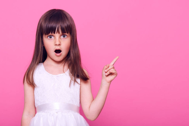 Surprised female child wearing beautiful white dress pointing aside with fore finger with widely opened mouth, sees something astonishing, copy space, isolated over pink background. - Foto, afbeelding