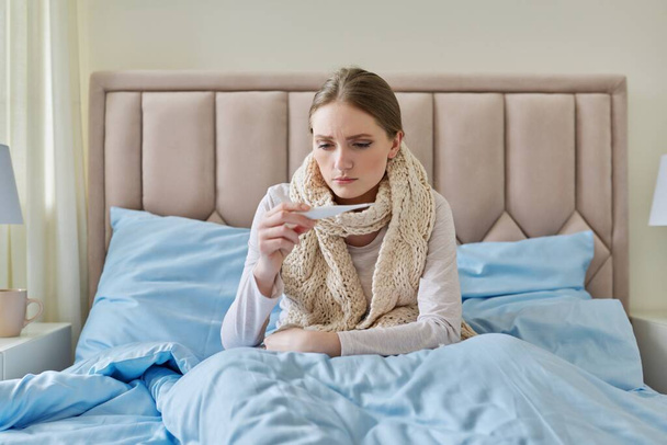 Sick young woman with thermometer in her hands, sitting at home in bed - Photo, Image