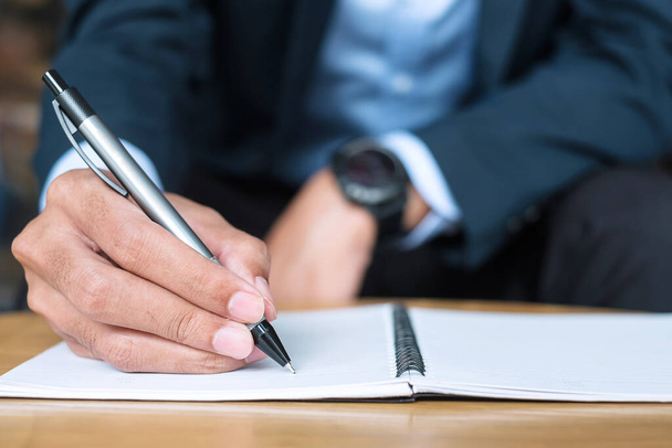 Biznesmen w garniturze piszący coś na notesie w biurze lub kawiarni, ręka człowieka trzymającego długopis z podpisem na papierowym raporcie. koncepcje biznesowe - Zdjęcie, obraz