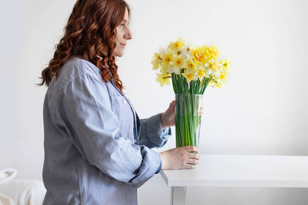 Schattig krullend haar meisje zet een transparante vaas met lentebloemen op een witte tafel in de kamer - Foto, afbeelding