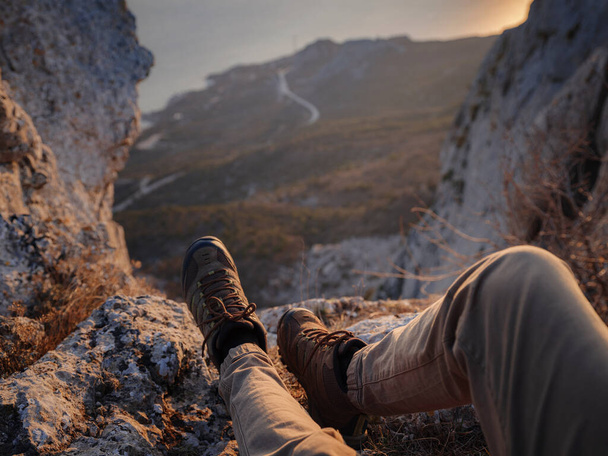 Закройте вид на мужские ноги в туристических ботинках. Человек турист сидит на краю скалы горы над морем - Фото, изображение