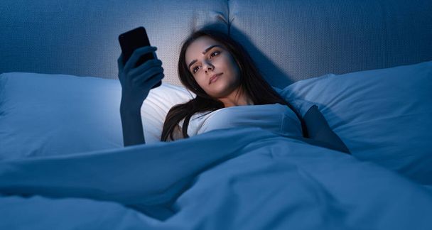 Young female with smartphone lying on bed - Valokuva, kuva