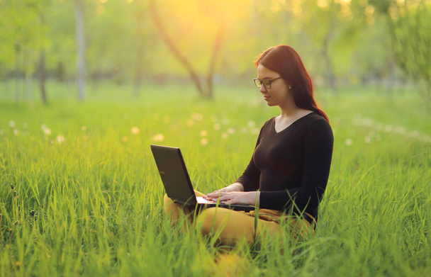 Cute smiling girl with laptop sits on green grass in the park and works remotely as a freelancer - Φωτογραφία, εικόνα