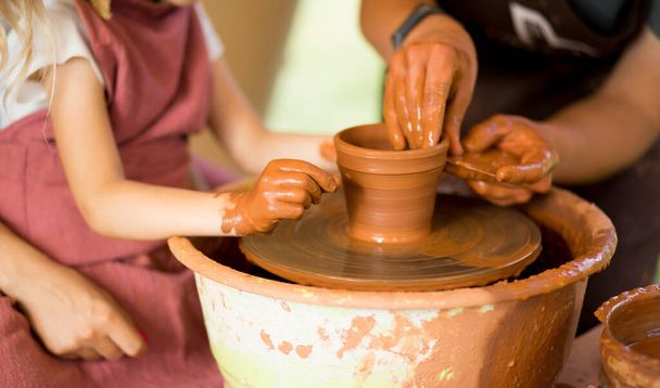 Clase magistral para niños. Las manos maestras enseñan al chiquitín, cómo hacer los platos de cerámica sobre el volante de alfarero. Artista trabaja con arcilla. Niña esculpir taza de arcilla - Foto, Imagen