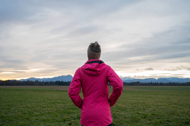 Вид сзади женщины в ярко-розовой куртке, стоящей на красивом лугу под вечерним небом, смотрящей вдаль. - Фото, изображение