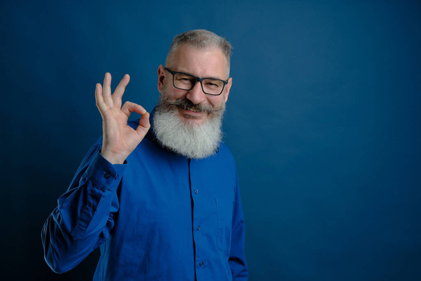 Retrato de hombre caucásico barbudo de pelo gris maduro vestido camisa azul muestra signo ok, fondo azul, enfoque selectivo - Foto, Imagen