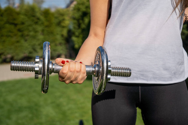 A sporty woman holding a dumbbell and working out in the park - Фото, изображение