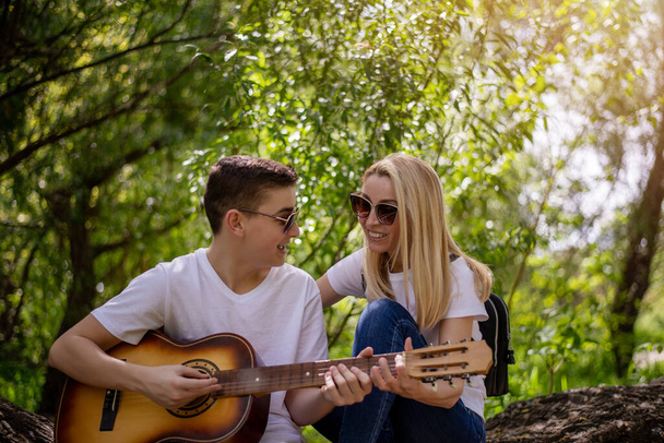 Chłopiec i jego matka grają i śpiewają na gitarze w letnim lesie. Mama siedzi na starym upadłym drzewie i cieszy się ze swojego syna. Ciesz się i odpoczywaj.. - Zdjęcie, obraz