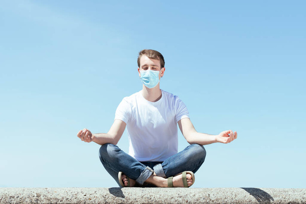 Медитация, йога и психическое здоровье во время Коронавируса. Красивый мужчина сидит в позе лотоса на улице и носит защитную маску. - Фото, изображение
