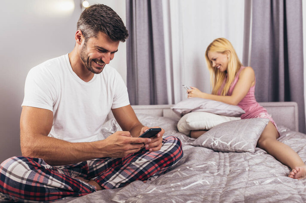 Young couple lying separate in a bed, man using phone. - Photo, image
