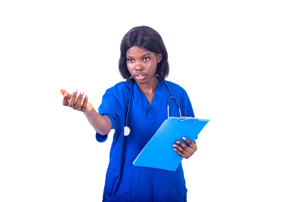 joven enfermera encantadora en bata de laboratorio de pie sobre fondo blanco con estetoscopio y carpeta haciendo gesto de la mano - Foto, imagen
