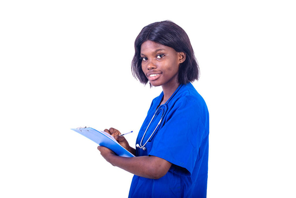 young charming nurse in lab coat standing on white background with stethoscope writing in folder and looking at camera smiling - Photo, Image