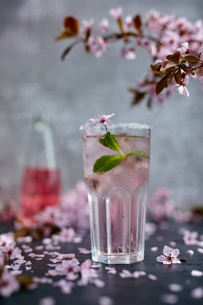 Tavaszi koktél. Egy pohár rózsaszín rózsa pezsgő, almabor vagy limonádé jéggel és mentával. Virág cseresznye ágak felett és szétszórt cseresznyevirág. Fekete-szürke háttér - Fotó, kép