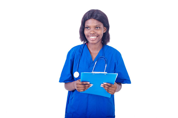 młoda urocza pielęgniarka w płaszczu laboratoryjnym stojąca na białym tle z folderem trzymającym stetoskop i patrząca na uśmiechniętą kamerę - Zdjęcie, obraz