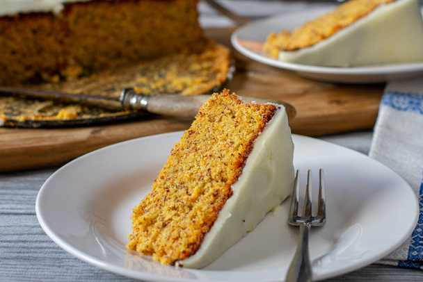 Крупный план кусочка свежей выпечки швейцарского морковного торта с миндалем и лимонной глазурью на белой тарелке с вилкой для торта подается на деревенском кухонном фоне. Готов поесть - Фото, изображение