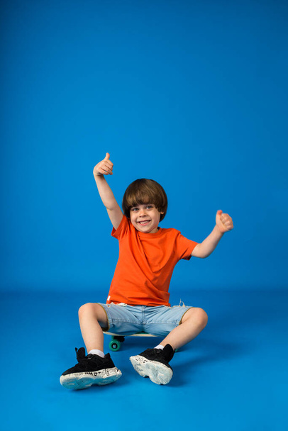 幸せな男の子幼児はスケートボードに座って、テキストのためのスペースを持つ青い背景に手を上げています - 写真・画像