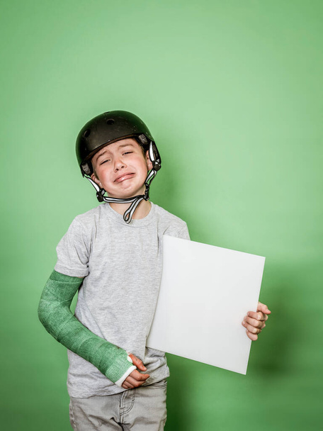 cool young schoolboy with broken arm and green plaster with black helmet holding white board in front of green background - Foto, imagen