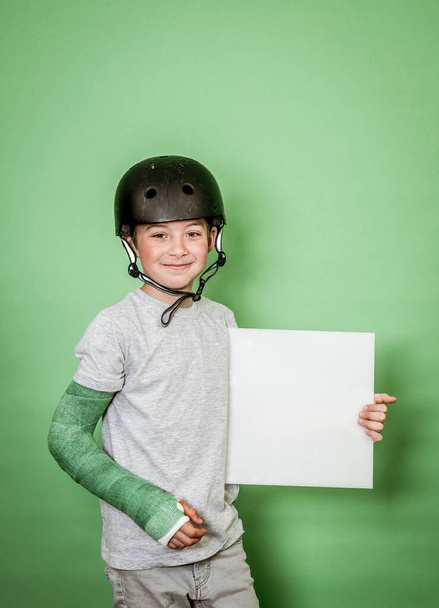 cool young schoolboy with broken arm and green plaster with black helmet holding white board in front of green background - Fotoğraf, Görsel