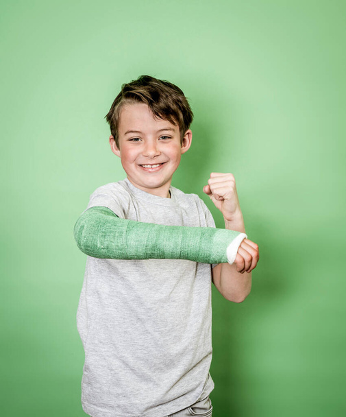 cool young schoolboy with broken arm and green arm plaster posing in front of green background - Foto, imagen