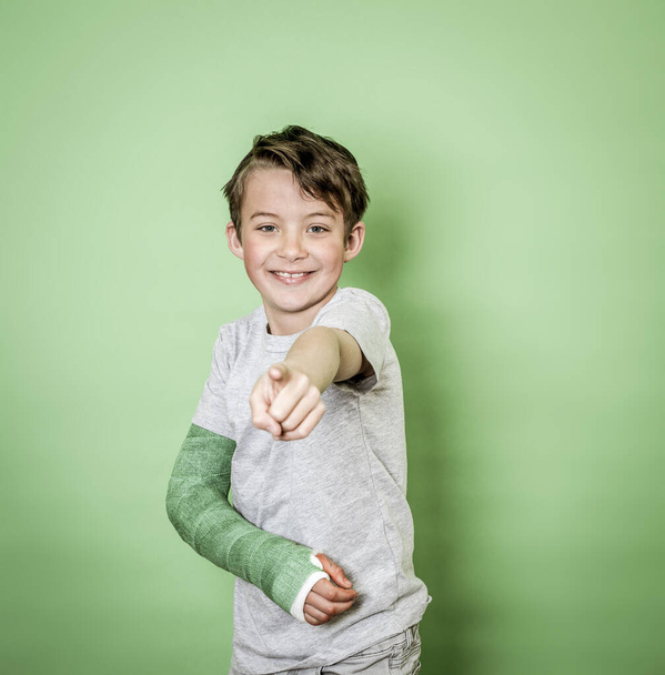 cool young schoolboy with broken arm and green arm plaster posing in front of green background - Фото, изображение
