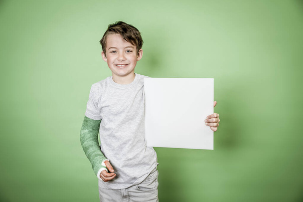 cool school boy with green hand plaster holding white board in front of green background - Photo, Image