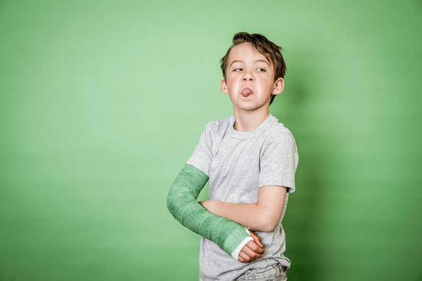 cool young schoolboy with broken arm and green plaster posing in front of green background - Foto, Imagem