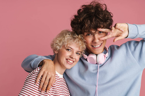 Happy young couple of teenagers standing isolated over pink background, wearing t-shirts, hugging, having fun - 写真・画像