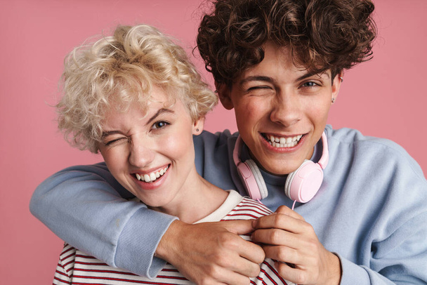 Happy young couple of teenagers standing isolated over pink background, wearing t-shirts, hugging, having fun - 写真・画像