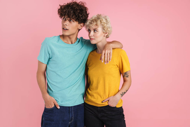 Attractive young couple of teenagers wearing t-shirts standing isolated over pink background, looking away - Foto, afbeelding