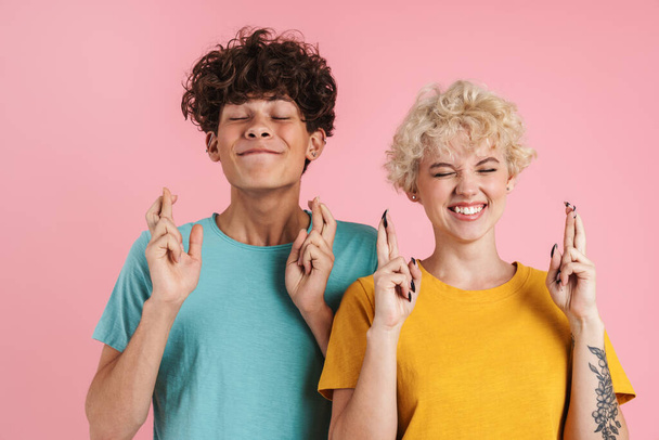 Happy couple holding fingers crossed for good luck isolated over pink background - Foto, afbeelding
