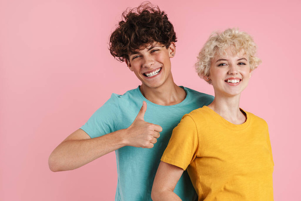 Happy young couple of teenagers standing isolated over pink background, wearing t-shirts, giving thumbs up - Fotografie, Obrázek