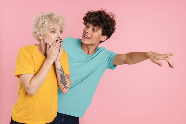 Portrait of an excited satisfied couple standing and pointing fingers away at copy space isolated over pink wall background - Фото, изображение
