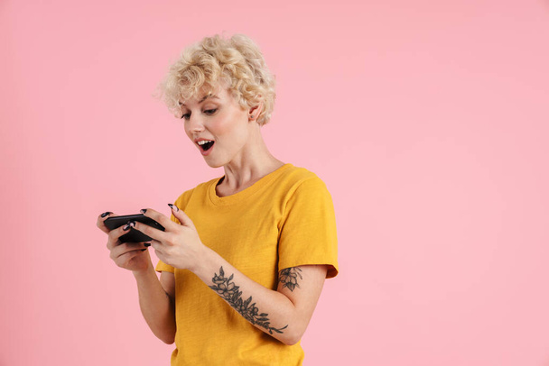 Portrait of a cheerful young blonde girl with curly blonde hair standing isolated over pink background, using mobile phone - Valokuva, kuva