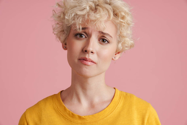 Portrait of a young upset blonde girl with curly blonde hair standing isolated over pink background - 写真・画像