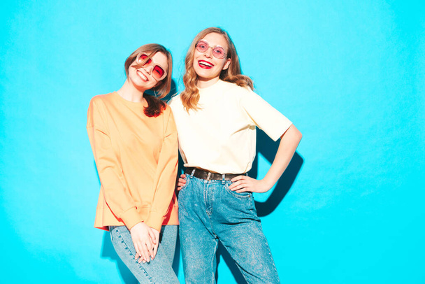 Two young beautiful smiling hipster female in trendy summer clothes. Sexy carefree women posing near blue wall in studio. Trendy and positive models having fun. Cheerful and happy - Valokuva, kuva