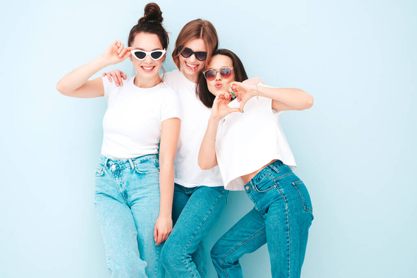 Three young beautiful smiling hipster female in trendy same summer white t-shirt and jeans clothes. Sexy carefree women posing near light blue wall in studio. Cheerful and positive models having fun - Foto, Imagem