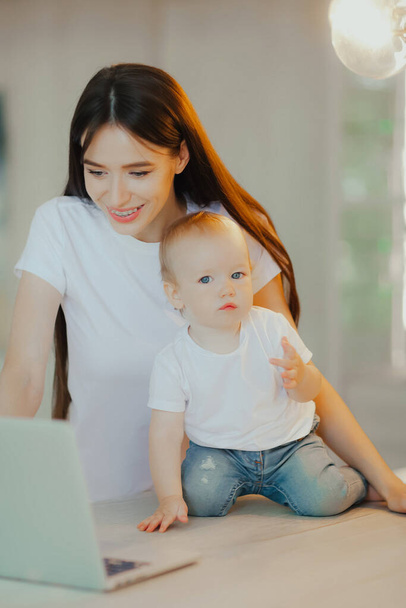 Little kid plays with laptop while mom looks after him. Baby care and technology - Zdjęcie, obraz