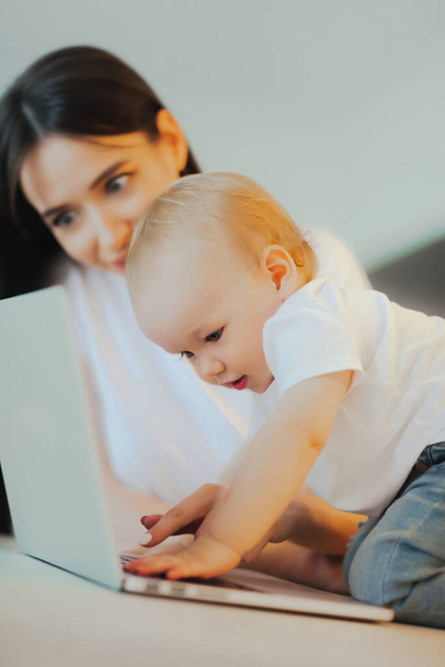 Kleines Kind spielt mit Laptop, während Mama sich um ihn kümmert. Babypflege und Technologie - Foto, Bild