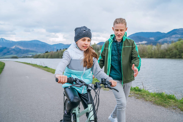  Happy outdoor childhood concept image. Brother carefully following his sister helping her and teaching doing first steps in bicycle riding.  - Zdjęcie, obraz