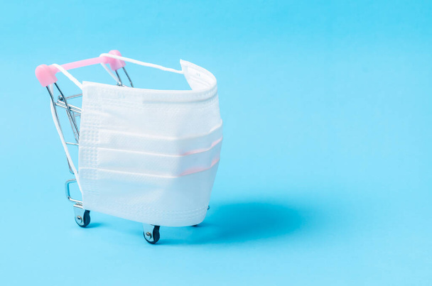 Disposable face mask medical and shopping cart on color background with empty space. Safe and online shopping on quarantine concepts during Covid - 19. - Фото, изображение