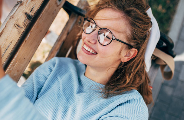 Gorgeous female in blue sweater, eyeglasses, smiling, and lying on the bench taking selfie on mobile phone. Happy woman takes a rest during online conversation on a smartphone outside. - Foto, Imagen