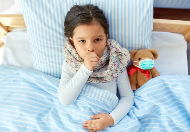 sick coughing girl with teddy bear lying in bed - Fotó, kép