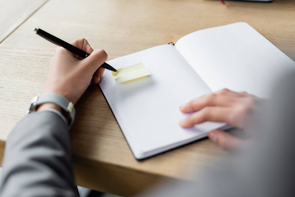 Gehakte weergave van wazig zakenvrouw houden pen in de buurt plakkerige notitie op notebook  - Foto, afbeelding
